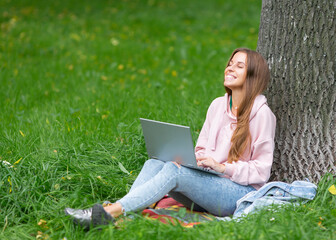 Happy laughing student girl having a video call, while sitting on the grass in city park