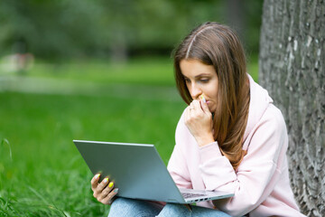 Pensive student girl having a video call, while sitting on the grass in campus park.