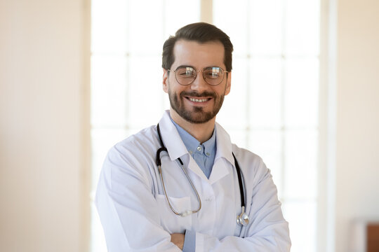 Head shot portrait smiling young man doctor wearing glasses and white coat with stethoscope looking at camera, confident successful therapist gp standing in office with arms crossed
