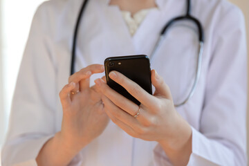Close up young woman doctor therapist gp wearing white uniform with stethoscope holding smartphone,...