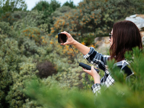 A young girl with dark hair, wearing glasses, takes photos of nature on her smartphone. The smartphone is charged from the power Bank. The concept of modern Hiking. a keepsake photo from the trip