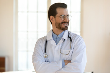 Smiling dreamy young man doctor wearing glasses and white uniform with stethoscope looking to aside, standing with arms crossed in office, confident successful therapist physician pondering future
