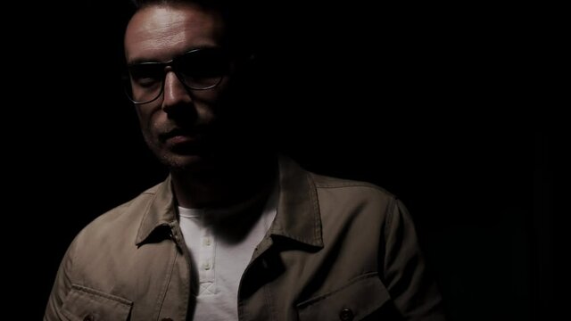 dramatic fashion model in beige jacket holding hand to chin and neck, looking up, rotating head, touching face and lips, standing in a shadow on black background