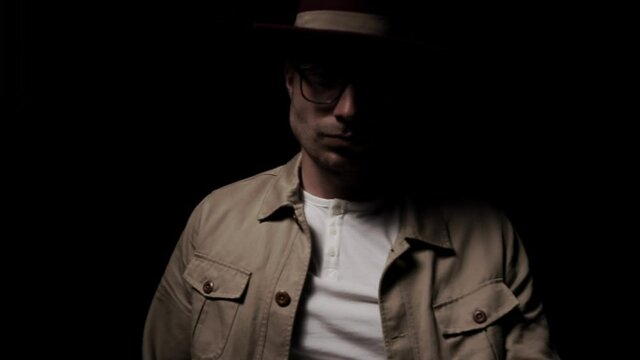mysterious young man fixing hat, sensually touching neck, looking to side and moving, arranging beige jacket, fixing glasses, walking in a shadow on black background