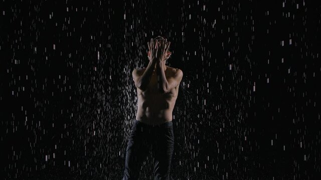 Wet young man dancer in jeans and with a naked torso performs contemporary dance in the rain and splashes of water. Male sensual contemp on black background. Slow motion. Close up.