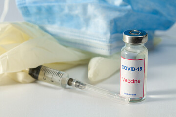 
In the foreground is a bottle of COVID-19 vaccine with the words "Made in Israel". In the background is a medical injection syringe, medical gloves and a medical mask out of focus.
