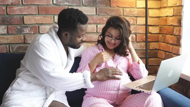 Handsome african man stroke wife's pregnant tummy while she is sitting with laptop, love and expecting baby concept.