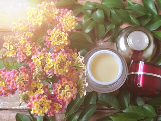 Facial cosmetic cream container,green leaves,colorful flowers on old wood plank.Rustic still life,soft dark tone,dimly light,with lens flare,free space for your text design or your products.