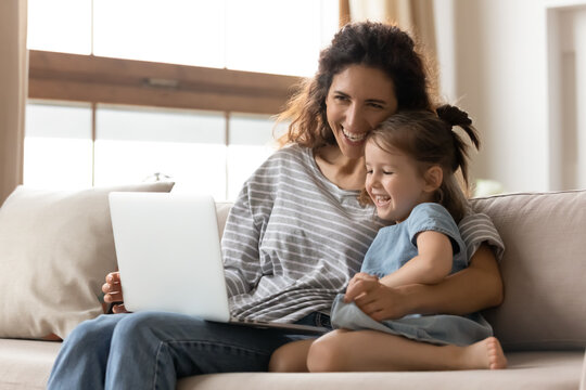 Attractive mother hug little daughter sitting on couch watching educational funny cartoons on pc laughing enjoy time together on weekend at home. Develop child and having fun using modern tech concept