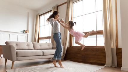 Fototapeta na wymiar Full length happy mother holds hands spinning carefree adorable little daughter listen music moving in modern interior light cozy living room. Active funny games, lively play have fun together at home