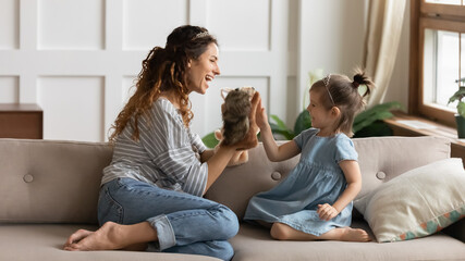 Mother and little daughter wear crowns sitting on sofa playing with hand puppet soft toys. Parent...