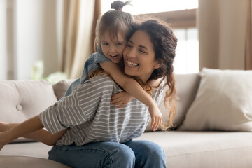 Mother piggybacks her little preschool cute daughter seated on couch enjoy funny active playtime together at home. Kid girl play with baby sitter in living room hug her from behind laughing having fun - obrazy, fototapety, plakaty