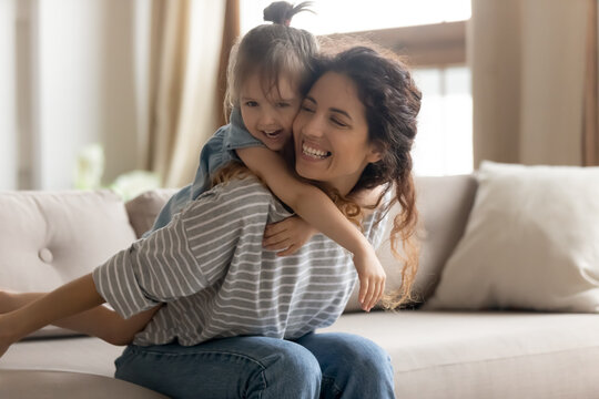 Mother piggybacks her little preschool cute daughter seated on couch enjoy funny active playtime together at home. Kid girl play with baby sitter in living room hug her from behind laughing having fun