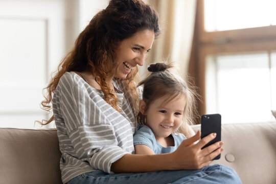 Mum and curious little daughter sit on couch hold cell phone watch cartoons use new funny app, during video call conference mom and kid girl laughing enjoy distant talk, parental control, secure usage