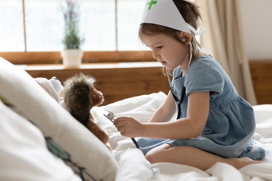 On bed focused little girl play nurse wear medical cap use stethoscope check listen heartbeat of soft hedgehog toy, loves cares about animals pretending to be doctor. Veterinary and healthcare concept