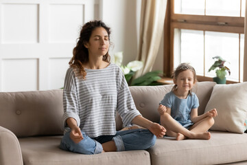 Serene young mother her little adorable daughter meditating breath fresh air sitting resting cross-legged on couch in living room. Healthy lifestyle yoga practice, parent teach kid of calmness concept