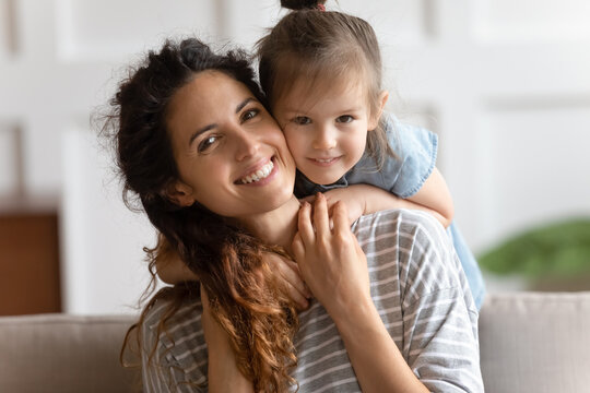 Happy motherhood, unconditional love, beautiful mother and little daughter portrait concept. Lovely kid girl hangs on mom neck, hugs her from behind piggybacking sit on couch smiling looking at camera