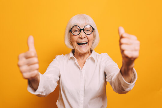 Image of old woman with white short hair and round glasses wearing white short, smiling and looking at camera. Blurred double thumb up sign on foreground. Woman isolated over orange background.