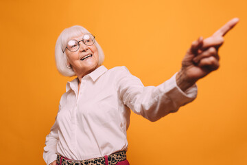 Elderly pretty gray-haired woman with round glasses wearing light blouse, standing isolated over orange background and pointing a finger at something.