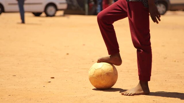 Medium close-up of young boy standing with his bare foot on soccer ball in dusty rural field of dirt before stepping back and kicking it on bright sunny day