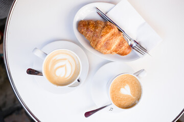 Two lattes coffee in white cups on a saucer and a croissant in a white plate, a fork and a knife on a summer terrace in a cafe. Girl with a phone in the background. Top view. flat lay