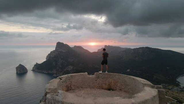 Aerial dolly out shot of a male filmmaker filming itself with a drone while standing on a watchtower at sunset