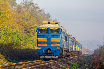 An old diesel locomotive with a commuter train arrives at a stopping point. Sunny autumn day.