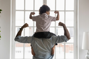 Back side view young african american father holding on shoulders little preschool funny kid son, showing biceps. Fit strong different generations family enjoying domestic activity, healthcare concept - obrazy, fototapety, plakaty