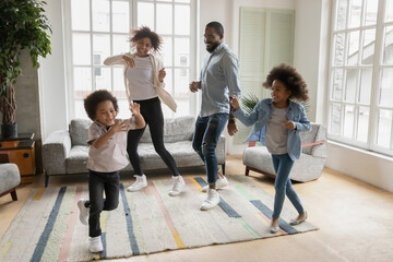 Full length overjoyed african ethnicity family having fun in living room, celebrating freedom. Happy mixed race young couple parents dancing to music with energetic funny biracial children siblings. - Powered by Adobe