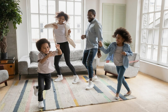 Full length overjoyed african ethnicity family having fun in living room, celebrating freedom. Happy mixed race young couple parents dancing to music with energetic funny biracial children siblings.