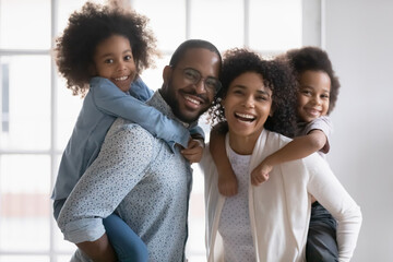 Portrait of happy biracial young couple parents giving piggyback ride to smiling adorable children siblings at home. Playful active family having fun with small kids indoors, looking at camera. - Powered by Adobe