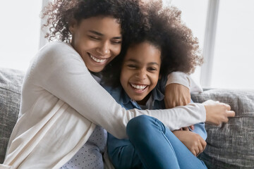 Loving young african ethnicity mother embracing laughing cute child daughter, having fun together on comfortable sofa at home. Affectionate caring biracial mum enjoying weekend with kid girl.
