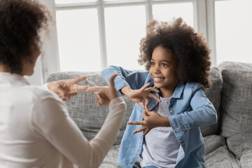 Smiling little african american girl with hearing loss practicing body language with professional female therapist at home, happy deaf kid communicating nonverbal with mum using signs and gestures. - Powered by Adobe