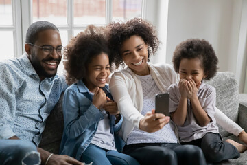 Happy young african american woman holding smartphone in hands, posing for selfie photo or recording funny video with playful laughing small adorable children siblings and biracial husband at home.