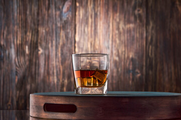 A square glass of whiskey on the rocks sits on a tray against an old wooden wall.