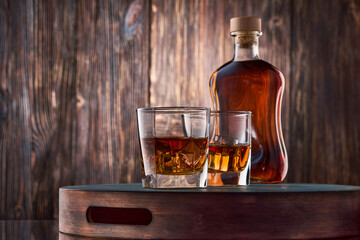 Two square glasses of iced whiskey and a full bottle of whiskey stand on a tray against an old wooden wall.