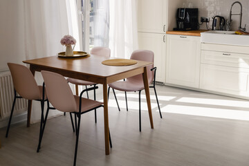 Fototapeta na wymiar Wooden dining table with four pink plastic chairs around it and a pink flower standing on it in a modern style kitchen on a sunny day