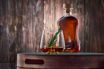 Fototapeta na wymiar A crystal glass of whiskey with a sprig of rosemary and a full bottle of whiskey stand on a tray against an old wooden wall.