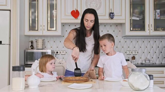 Family of mother and two kids cutting raspberry pie in kitchen. Pretty mother with little son and daughter cooking cake at home. 4K, UHD