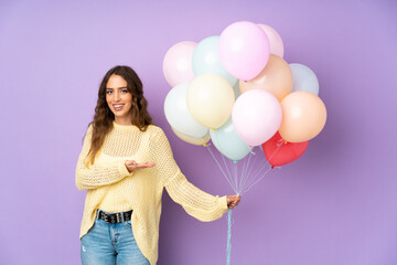 Young woman catching many balloons over isolated on purple background extending hands to the side for inviting to come