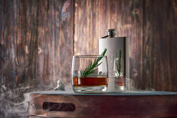 A round glass of whisky with a sprig of rosemary and a metal flask, shrouded in clouds of smoke,...