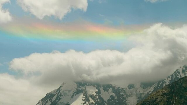 Mountain peaks covered with snow woth thick fluffy clouds and a rainbow above them. Nice view of russian nature.