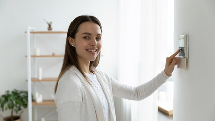 Fototapeta na wymiar Happy young female client looking at camera, pressing buttons on smart house system. Smiling millennial woman using indoors temperature controller or air conditioning, setting alarm password.