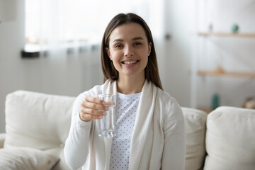 Portrait of happy beautiful young woman sitting on comfortable sofa, holding glass of fresh pure water. Smiling millennial girl looking at camera, offering drinking aqua every day, healthcare habit.