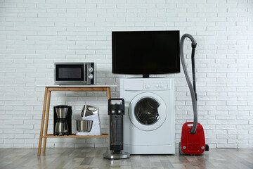 Modern vacuum cleaner and different household appliances near white brick wall indoors