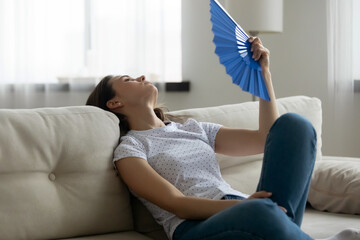Exhausted young woman waving paper fan, suffering from high indoors temperature. Tired millennial...