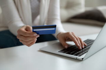 Close up young woman holding plastic banking credit debit card, involved in online shopping on computer at home. Female client entering payment information cvv code confirming purchase indoors.