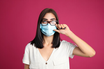 Young woman wiping foggy glasses caused by wearing disposable mask on pink background. Protective...