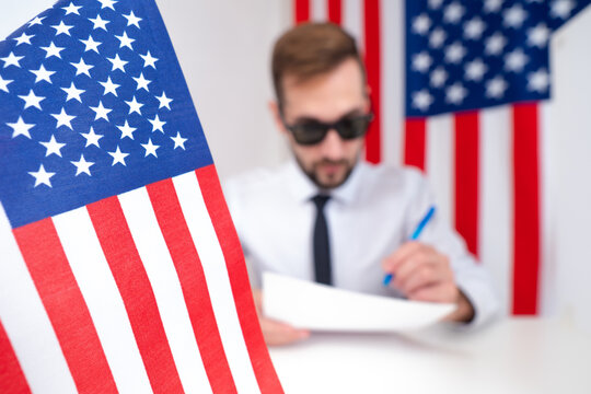 A man with black glasses writes against the background of the American flag. The work of American intelligence agencies. American spy. An official in the USA coordinates the documents.