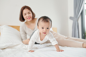 Fototapeta na wymiar Young Asian mother resting on bed and looking at her adorable crawling baby